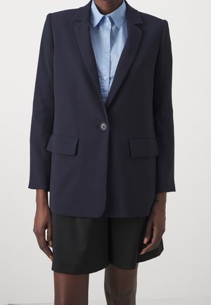 Blazer bleu marine foncé à un bouton porté sur une chemise bleu clair à col et un short noir ample, mannequin debout sur un fond uni.