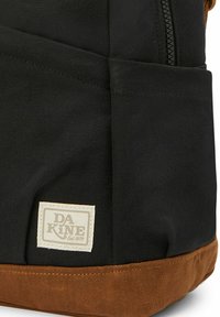 Zwarte stoffen rugzak met een bruine bodem, voorzien van een gestikte logopatch. Textuurmateriaal met zichtbare naden en ritsdetails.