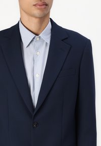 Marineblazer mit einem strukturierten Design, Ein-Knopf-Verschluss und einem hellblauen Hemd darunter. Glatter Stoff mit maßgeschneidertem Schnitt.