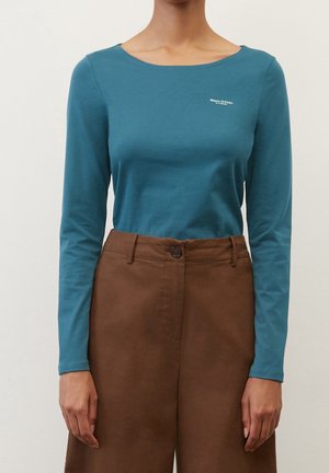 Person trägt ein türkisfarbenes Langarmshirt mit dem "Marc O'Polo"-Logo und hoch taillierte braune Hosen mit Knopfverschluss.