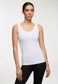 Donna in piedi, con indosso un top bianco aderente senza maniche e leggings neri, con le braccia rilassate lungo i fianchi contro uno sfondo neutro.