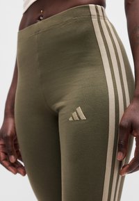 Κοντινή λήψη ατόμου που φοράει ελαστικό παντελόνι ελιάς με μπεζ ρίγες και λογότυπο Adidas, δείχνοντας το μέσο σώμα και τα χέρια να ακουμπούν στα πλάγια.