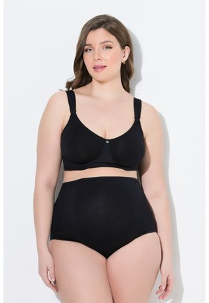 Mujer de piel clara y cabello castaño, vestida con un sujetador negro y ropa interior de tiro alto, de pie frente a un fondo blanco.