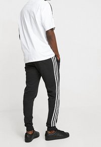 Man som bär svarta träningsbyxor med vita ränder, vit t-shirt och svarta sneakers, står med händerna i fickorna mot en enfärgad bakgrund.