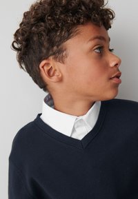 Tmavomodrý sveter s výstrihom do V nosený cez bielu košeľu s golierom. Košeľa má vzorovaný golier viditeľný na výstrihu.