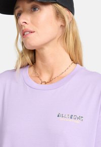 Lyse lilla bomulds T-shirt med rund halsudskæring og et lille guldtrykt "BILLABONG" logo på forsiden. Kædetrøje synlig.