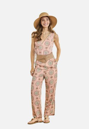 Mujer con un mono sin mangas rosa con estampado floral, sombrero de ala ancha, cinturón de cintura con tachuelas marrón, sandalias y un collar de conchas, sonriendo con una mano en el bolsillo.