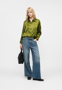 Camisa verde de cetim, abotoada, com mangas longas, combinada com calças de ganga azuis de perna larga. Uma mala e sapatos pretos completam o look.