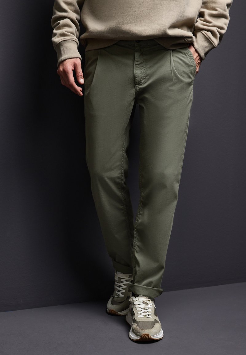 Olivově zelené chinos s mírně texturovaným povrchem, zúženými nohavicemi, předními záhyby a ohrnutými manžetami, doplněné světlými béžovými teniskami.