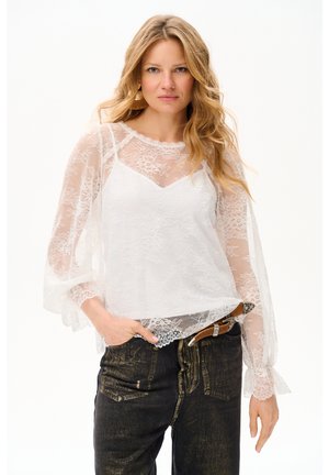 Kobieta o falowanych blond włosach, ubrana w przezroczystą białą koronkową bluzkę na białym podkoszulku i ciemnych dżinsach z brązowym paskiem, stoi z jedną ręką w kieszeni.