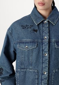 Chaqueta de mezclilla azul con cierre de botones, dos bolsillos frontales y texto bordado a lo largo del cuello, además de un diseño en la manga.