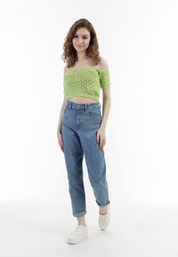 Pletený crop top v jasně zelené barvě s odkrytými rameny a strukturovaným vzorem, doplněný o světle modré džíny s vysokým pasem a bílé tenisky.
