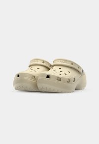 Sabots Crocs beiges avec un design perforé, une large sangle et une semelle texturée. Fabriqués en matériau léger avec une forme arrondie.