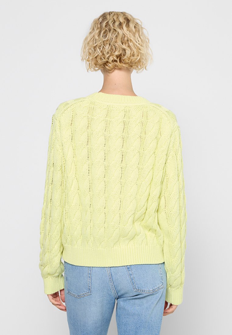 Pull en tricot jaune clair avec un motif en câble, ourlets et poignets côtelés, avec une coupe décontractée. Porté avec un jean bleu taille haute.
