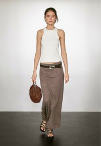 Witte mouwloze top, lange bruine rok, zwarte riem met zilveren gesp, bruine gevlochten tas en zwarte vetersandalen. Eenvoudige, lange silhouet.