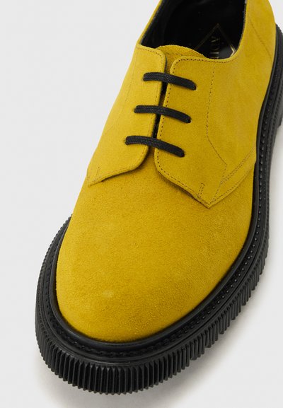 Chaussure montante en daim jaune avec semelle épaisse noire à rainures sur fond blanc.