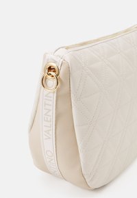Prošívaná béžová kožená taška se zlatými doplňky, s bílým brandovaným popruhem a zipovým uzávěrem, navržená s kulatým tvarem.