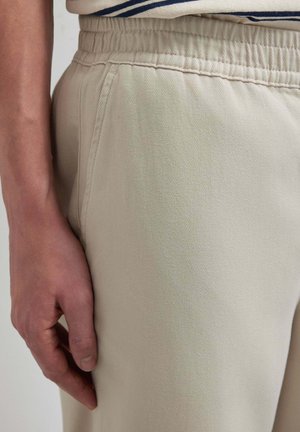 Linke Hand ruht neben beige-farbenen Hosen mit elastischem Bund, sichtbarer Seitentasche und einem gestreiften Hemd, das eingesteckt ist.