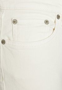 Bílé džíny vyrobené z denimového materiálu, které mají přední kapsu se stříbrnými detaily a jemnou texturou. Tkanina má mírný vzor keprového tkaní.