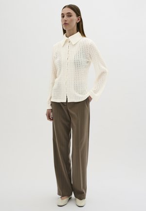 Camicia a maglia color crema con bottoni e colletto a punta, caratterizzata da un motivo testurizzato, abbinata a pantaloni marroni a righe sottili e scarpe chiare.