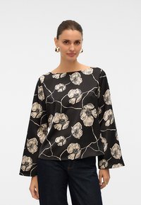 Mujer con el cabello recogido, que lleva pendientes de aro dorados, una blusa negra de manga larga con estampados florales beige y vaqueros azul oscuro.