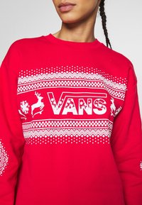 Sweat-shirt rouge avec des imprimés graphiques blancs, comprenant le logo "VANS", un renne et des motifs géométriques. Tissu doux avec un col rond.