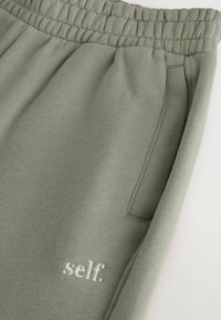 Pantaloni sportivi verde chiaro realizzati in tessuto morbido, con vita elasticizzata, tasca laterale e logo "self." ricamato in bianco.
