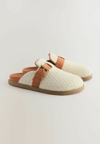 Beige gestrickte Slipper mit einer strukturierten Oberfläche, ausgestattet mit einem braunen Akzentriemen und einer goldenen Schnalle. Gummisohle für guten Halt.