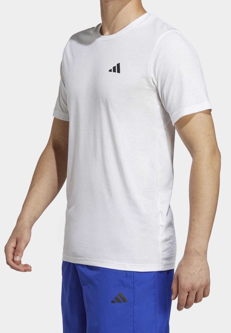 Muž v bílém tričku s krátkým rukávem a modrých šortkách, oboje s malým černým logem se třemi pruhy na hrudníku a stehně.