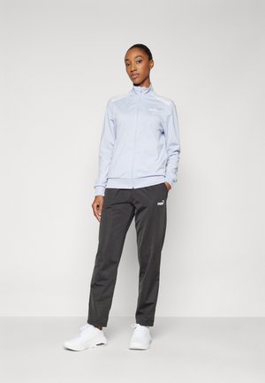 Jachetă cu fermoar de culoare albastru deschis, cu guler înalt, și pantaloni gri, având logo-uri. Pantofii sport albi completează ținuta.