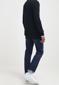 Svart stickad tröja med långa ärmar, tillsammans med mörkblå jeans. Skorna är vita sneakers och jeansens benslut är uppvikta.