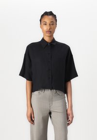 Camicetta nera cropped con bottoni, maniche corte e ampie, caratterizzata da una trama liscia e un collo classico. Abbinata a pantaloni dritti grigio chiaro.