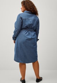 Abito camicia in denim di colore blu medio. Presenta maniche lunghe, vita con cintura e orlo curvo. Tessuto testurizzato con dettagli di cucitura visibili.