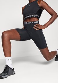 Atleta w czarnym stroju sportowym Nike Pro wykonujący rozciąganie w wykroku, w czarnych i szarych butach do biegania oraz białych skarpetkach Nike.