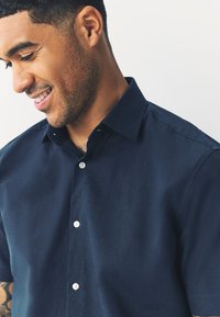 Camicia blu navy a maniche corte realizzata in tessuto dalla texture liscia. Presenta un colletto con bottoni e due bottoni bianchi sulla patta.