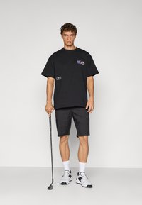 Svarte oversized t-skjorte med lomme og logo, kombinert med svarte shorts. Modellen holder en golfklubbe og har på seg hvite treningssko og sokker.