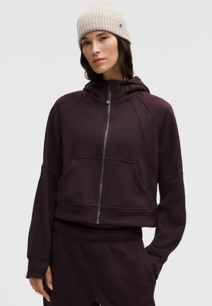 Rövidített zipzáros kapucnis pulóver mély lila anyagból, elülső zsebekkel és kapucnival. Hozzá illő nadrággal párosítva, laza fazonnal és bordázott mandzsettákkal.