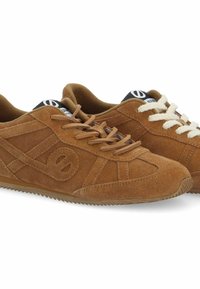 Baskets en suède brun avec des lacets contrastés blancs et bruns, des bords texturés et un logo distinctif sur un onglet noir au niveau de la languette.