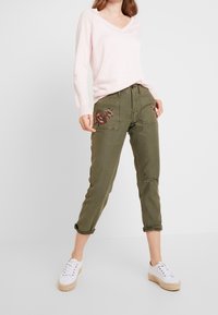 Světle růžový svetr s výstřihem do V zkombinovaný s olivově zelenými zkrácenými kalhotami s květinovou výšivkou a vybledlými akcenty, doplněný bílými teniskami.