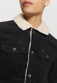 Personne portant une veste en velours côtelé noire avec un col sherpa crème et des boutons-pression métalliques, montrant la partie supérieure de la poitrine et du cou.