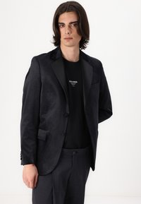 Chaqueta de esmoquin negra con solapas de satén, ajuste entallado, cierre de un botón y bolsillos laterales. Llevada sobre una camiseta negra y pantalones grises.