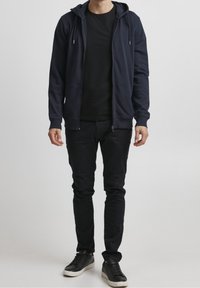 Sweat à capuche zippé bleu marine avec cordons, associé à un t-shirt noir, un pantalon noir slim et des baskets noires à semelles blanches. Tissu doux.