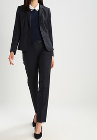 Svart skräddarsydd kostym med en åtsittande kavaj och raka byxor, lager på lager över en marinblå tröja och vit blus med krage. Klackskor.