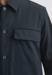 Dunkelblaues Hemdjackett aus leichtem Stoff, mit einer Brusttasche, einem klassischen Kragen und glatter Textur mit kontrastierenden Nähten.
