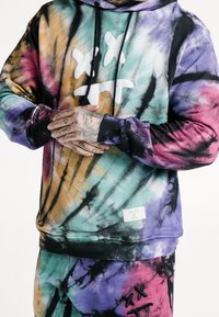 Dres z kapturem w kolorowe wzory tie-dye (fioletowy, błękitny, pomarańczowy). Posiada biały graficzny wzór, czarne akcenty oraz metkę z logo marki na dolnym brzegu.