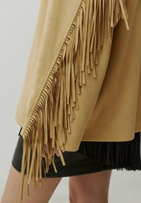 Tan suède poncho met lange franjes aan één zijde. Glad textuur met een gedrapeerd ontwerp, gecombineerd met een zwarte geplooide rok eronder.