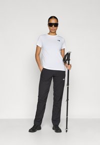 Λευκή κοντομάνικη μπλούζα με μαύρο λογότυπο, μαύρο ελαφρύ παντελόνι πεζοπορίας, μαύρα παπούτσια, κρατώντας μπαστούνι πεζοπορίας, φορώντας μεγάλα σκουρόχρωμα γυαλιά ηλίου.