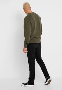 Man with shaved head wearing an olive green sweatshirt, black jeans, and black sneakers standing against a plain white background.