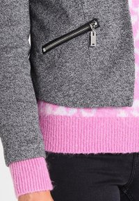 Šedá strukturovaná bunda s krátkým střihem, obsahující kapsu na zip a růžové vroubkové manžety, navrstvená přes růžový vzorovaný svetr.