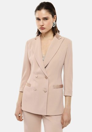 Donna che indossa un blazer doppiopetto beige con pantaloni abbinati e grandi orecchini d'argento su uno sfondo semplice.